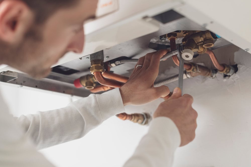 A person in a white long-sleeve shirt is using a wrench to work on plumbing pipes, focusing on tightening a fitting under a sink or perhaps a boiler. The image highlights their hands and the HVAC Repair Harford County components they are adjusting.