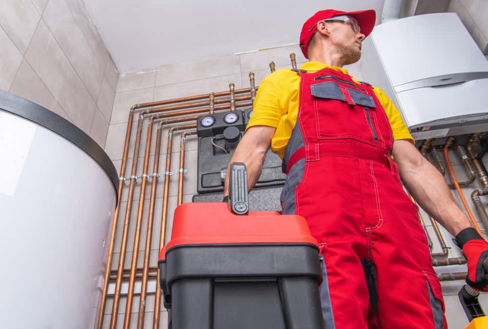 A worker dressed in red overalls and a cap, holding a toolbox, stands in a room with various plumbing pipes and a large white water heater or boiler. The individual looks towards the right, with copper pipes and heating equipment visible in the background, reminiscent of an HVAC company Harford County technician at work.