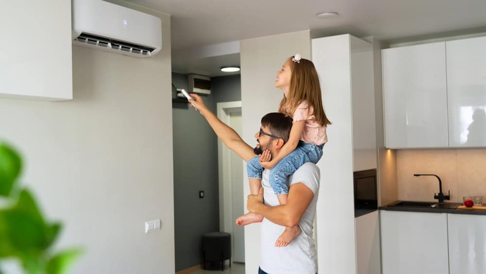 A man is carrying a young girl on his shoulders as she uses a remote control to operate their new air conditioner. They are inside a modern kitchen with white cabinets. The man is smiling, and the girl appears focused on the task, showcasing just how smoothly an ac repair from an HVAC company in Harford County can go.