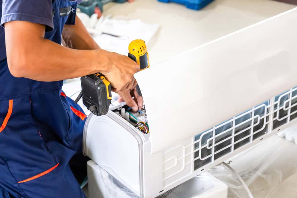 A technician in a blue uniform repairs an air conditioning unit while holding a screwdriver. The cover of the air conditioner is lifted, exposing its internal components. The scene suggests an HVAC Repair Harford County task in progress within a domestic or commercial setting.