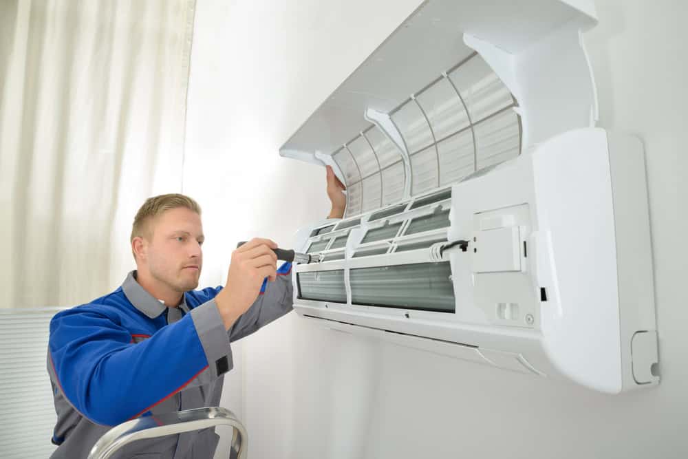 A technician wearing a gray and blue uniform is standing on a ladder, using a screwdriver to repair or install a wall-mounted air conditioning unit. The front cover of the unit is open, revealing the internal components—a typical scene during HVAC Repair in Harford County.