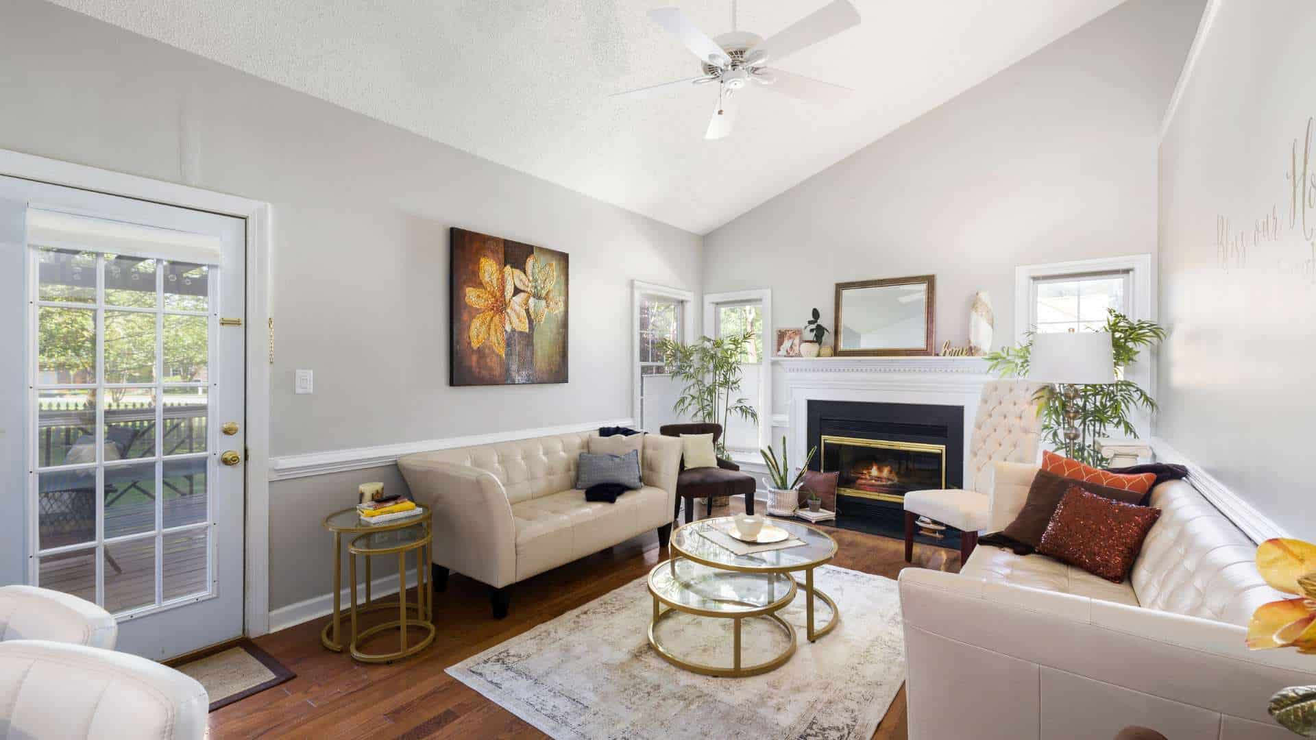 A cozy living room features white walls, a white ceiling fan from a reputable HVAC company in Harford County, and wooden flooring. It has white sofas, a glass coffee table, and a rug. There is a fireplace with a mirror and decor on the mantel, a large painting on the wall, and two windows letting in natural light.