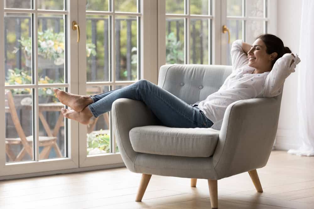 A woman is relaxing in a light gray armchair with her legs up on the armrest and her hands behind her head. She is smiling and looking out large windows that show a view of an outdoor patio with plants, appreciating the comfort provided by an ac repair Harford County service. The room is bright and airy.